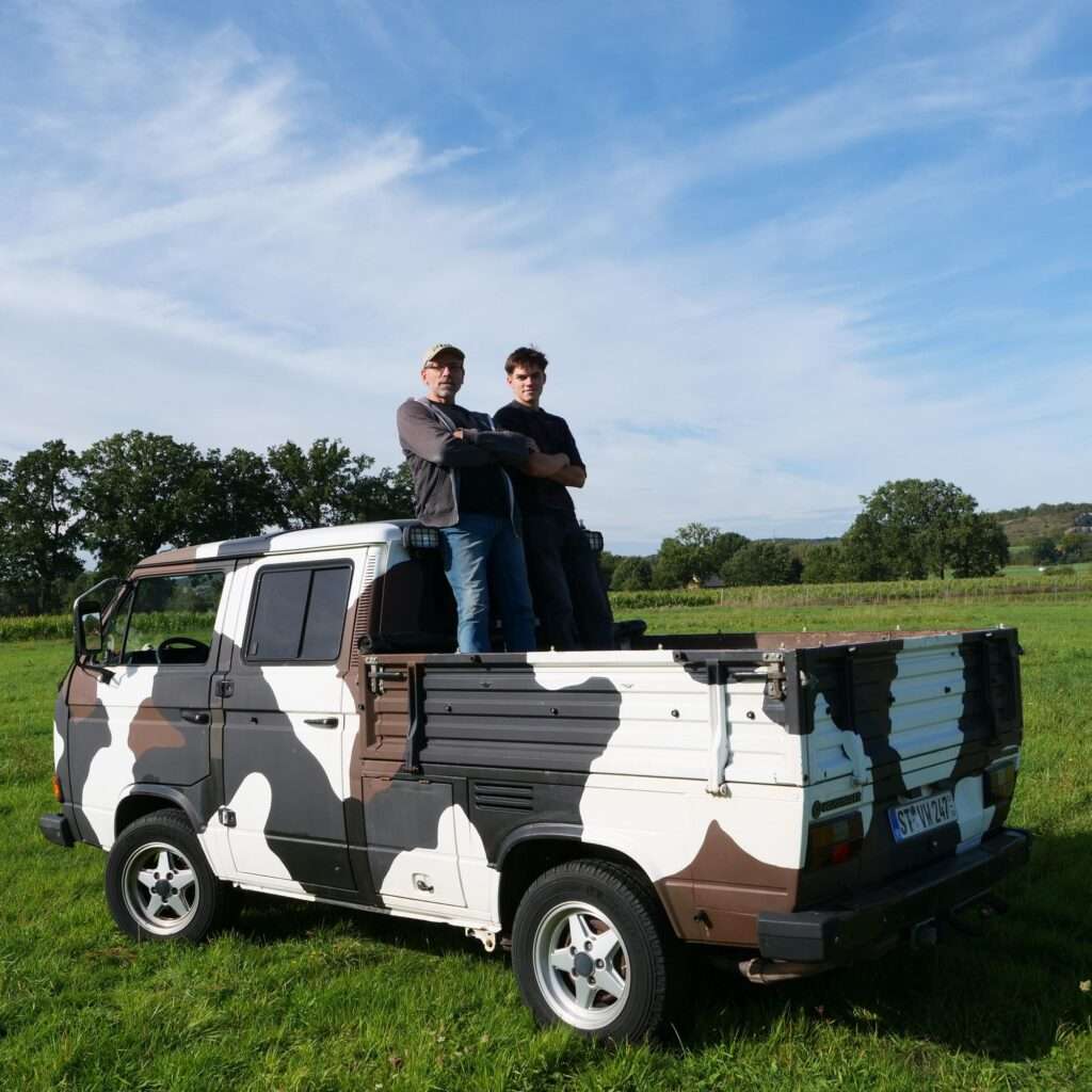 Vater und Sohn stehen auf der Ladefläche des kuhgefleckten VW T3 Doka